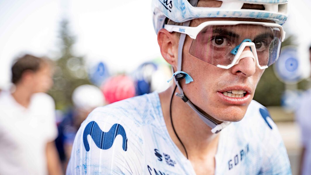 Close-up of a Movistar rider wearing the white young rider jersey at La Vuelta España, focused and determined during a stage, with blurred spectators in the background.