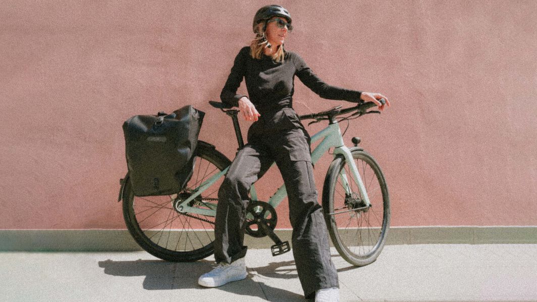 How to choose the right city or touring bike: Look for models with versatile mounting options for racks and accessories. Woman posing with a Canyon commuter bike in front of a minimalist pink wall, wearing casual black outfit and helmet